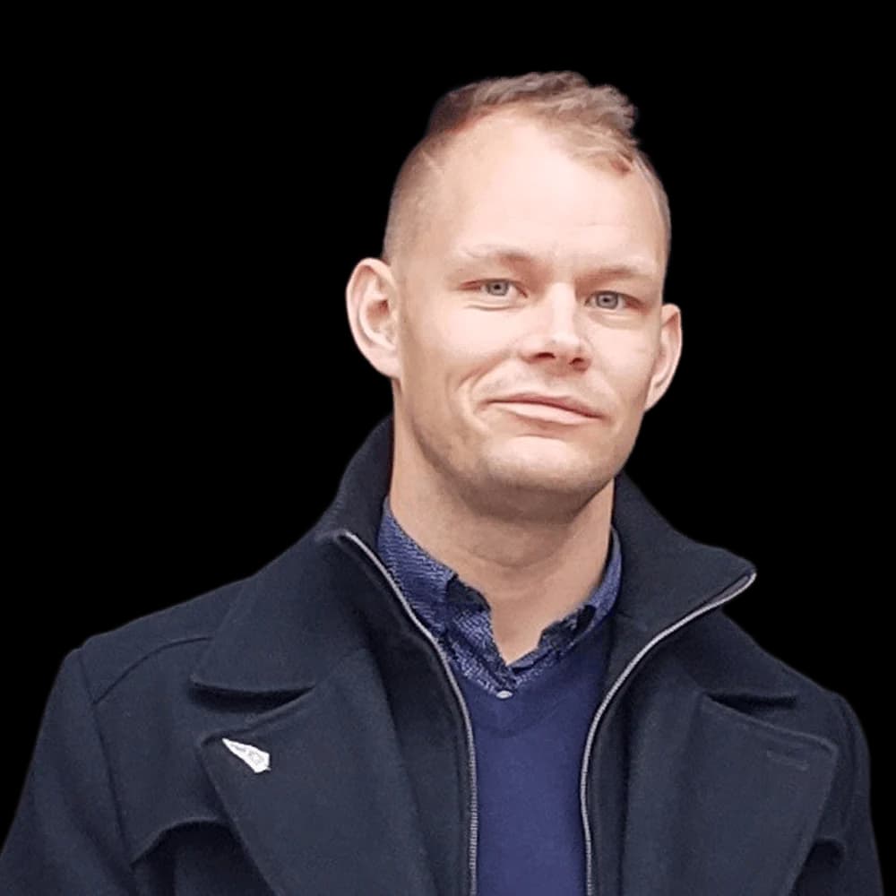 Fair-skinned man with short blonde hair and blue eyes wearing a dark blue coat.